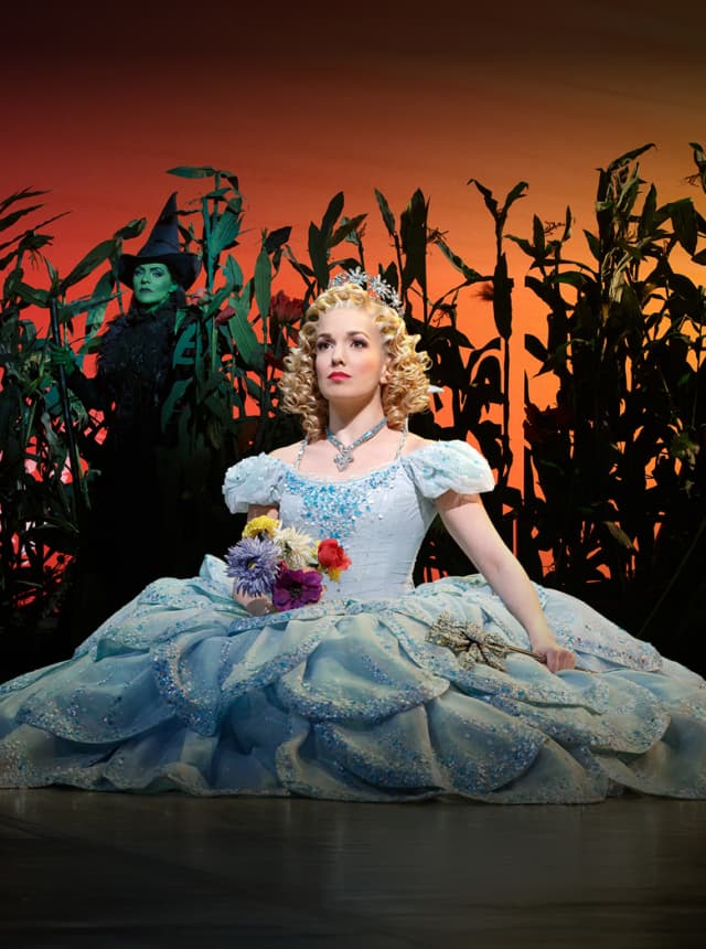 Glinda in a blue gown with Elphaba behind her in a cornfield at sunset.
